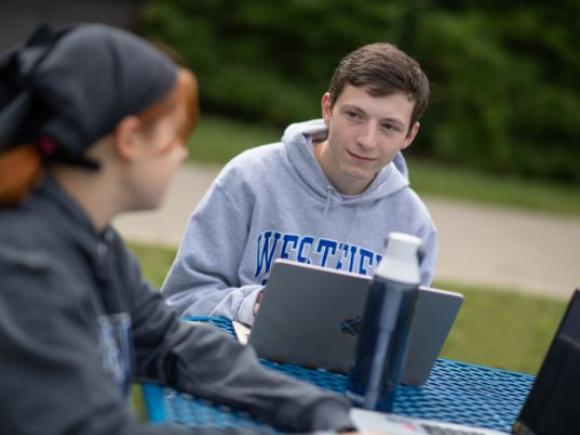 Two students work on their laptops while seated at a table outside on the 91ɫ campus.