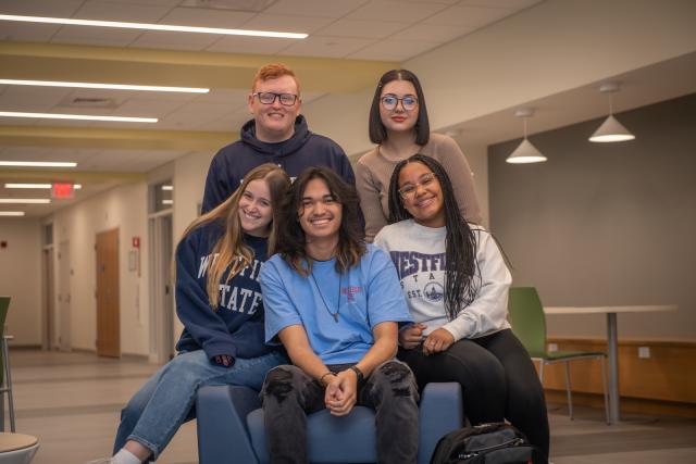 Five students wearing 91ɫ State sweatshirts smiling in Parenzo Hall. Two standing three sitting.