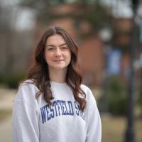 Paige Freeman, a senior at the University. She is outside, near the Ely building. She has long, brown hair and a gray sweatshirt which says "91ɫ State" in blue lettering. Blurred out trees, grass, and the sidewalk are in the background.